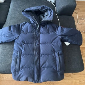 Mörkblå dunjacka från Canada Goose - Säljer en mörkblå dunjacka från Canada Goose med huva och klassisk logga på ärmen. Jackan har dragkedja framtill, två fickor och quiltad design. Perfekt för kalla vinterdagar och riktigt snygg streetstil. Kvitto skickas privat då de är mail ! 10 lax ny 