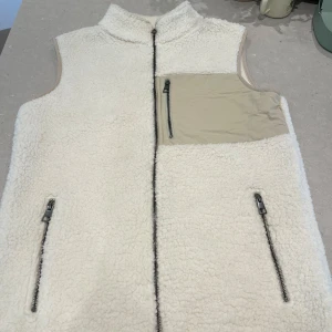 Vit fleeceväst med beige ficka - Säljer en vit fleeceväst med hel dragkedja framtill. Västen har två fickor med dragkedja och en beige bröstficka med dragkedja. Materialet är mjukt och fluffigt, perfekt för lager-på-lager. Enkel och clean design.