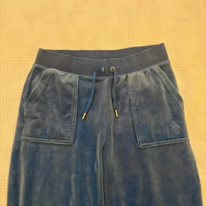Blå velourbyxor Juicy Couture S - Mysiga blå mjukisbyxor från Juicy Couture i velour med bred resår i midjan och snörning. Stora fickor fram och bak, varav bakfickorna har lock och tryckknappar. Broderad JC-logga framtill. Perfekta för en chill och trendig look.