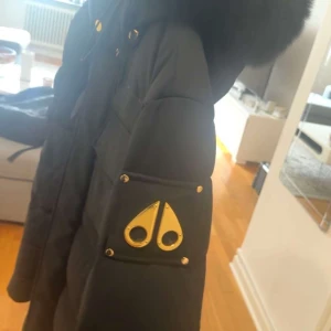 Svart dunjacka Moose Knuckles  - Säljer en svart dunjacka från Moose Knuckles med lyxiga gulddetaljer och stor pälskantad huva. Jackan har flera stora fickor framtill, dragkedja och tryckknappar, samt ett glansigt silverfoder på insidan. Perfekt för kalla dagar och riktigt snygg streetstil.