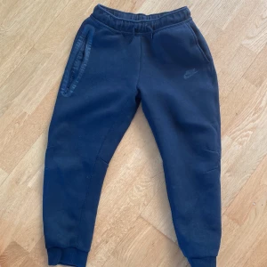 Svarta mjukisbyxor Nike tech  - Snygga svarta Nike tech byxor med resår och snörning i midjan. Byxorna har ribbade muddar vid bensluten och praktiska fickor med dragkedja. Perfekta för chill eller träning, med diskret Nike-logga på låret.