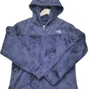 Blue fleece Jacka från The North Face women - Mysig blue teddyjacka från The North Face med huva och hel dragkedja framtill. Jackan har fluffig, mjuk yta i fleece och förstärkta partier över axlar och huva. Perfekt för kyliga dagar när du vill ha både stil och komfort.