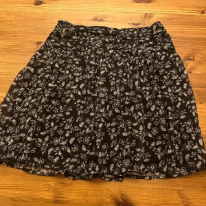 Svart blommig kortkjol från BikBok - Supersöt svart kortkjol från BikBok med vitt blommönster. Kjolen har en smockad midja som ger en skön passform och är gjord i ett lätt och luftigt material. Perfekt att styla med en enkel topp och sneakers för en chill look.