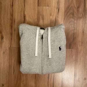 Grå hoodie från Polo Ralph Lauren - Snygg grå hoodie från Polo Ralph Lauren med dragkedja, vita snören och klassisk logga på bröstet. Tröjan har huva, känguruficka och är tillverkad i mjuk bomullsmix. Perfekt för chill dagar eller när du vill ha en clean look.