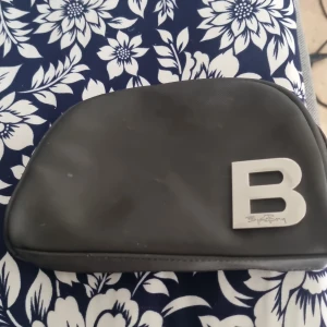 Svart plånbok Björn Borg med B-detalj - Svart plånbok från Björn Borg med stilren design och stor silverfärgad B-detalj framtill. Plånboken har dragkedja upptill och ett extra fack med dragkedja på baksidan. Tillverkad i syntetmaterial och perfekt för att hålla ordning på kort och småsaker.