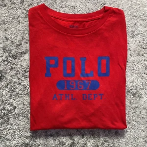 Röd ralph lauren T-shirt - Röd t-shirt från Polo Ralph Lauren med blått tryck framtill där det står 'POLO 1967 ATHL. DEPT.'. Klassisk rund hals och normal passform. Tillverkad i mjuk bomull, perfekt för en avslappnad stil.