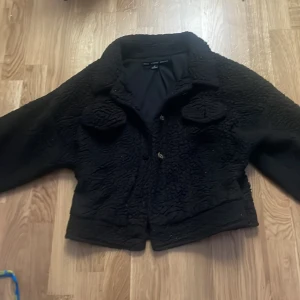 Svart teddyjacka från HYFVE - Mysig svart teddyjacka från HYFVE i storlek M. Jackan har två stora bröstfickor med lock, klassisk krage och knäppning framtill. Perfekt för dig som gillar en cool och avslappnad stil med lite extra fluff.