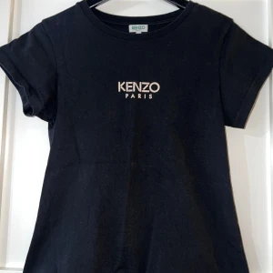 Svart Kenzo t-shirt - Svart t-shirt från Kenzo med klassisk rund hals och vit Kenzo Paris-logga tryckt på bröstet. T-shirten är kortärmad och har en enkel, stilren design som passar perfekt till jeans eller shorts. Tillverkad i mjuk bomull för skön känsla hela dagen. I mycket bra skick och knappt använd. 