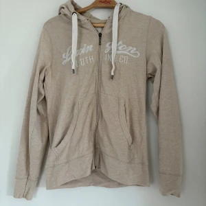 Beige hoodie från Lexington XS - Beige hoodie från Lexington i 100% bomull med broderad vit text på bröstet. Tröjan har huva med vita snören, dragkedja framtill och två stora fickor. Perfekt för en chill och avslappnad stil. Liten fläck finns 