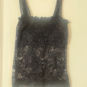 Svart spetslinne med blommönster - Säljer ett svart linne med breda axelband och vacker blommig spets över hela toppen. Snyggt genombrutet material som ger en cool och feminin vibe. Perfekt att styla lager på lager eller som den är till jeans eller kjol.