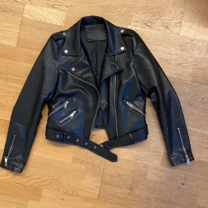 Skinnjacka - Svart skinnjacka med silvriga dragkedjor, axelklaffar och bälte i midjan. Jackan har flera fickor med dekorations dragkedjor, långa ärmar med dragkedjor vid ärmsluten. Perfekt för dig som vill ha en edgy och tidlös look.