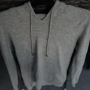 Grå stickad hoodie med snörning - Mysig grå stickad hoodie med snörning i halsen och lång ärm. Tröjan har en enkel och stilren design med ribbad kant vid halsen. Perfekt för dig som gillar en avslappnad och trendig look. Tröjan är i nyskick knappt använd. Storleken är M och tröjan är i slim fit. Skick 10/10