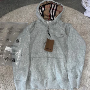Grå hoodie från Burberry med dragkedja - Snygg ljusgrå hoodie från Burberry med klassiskt rutigt foder i huvan. Tröjan har dragkedja framtill, snörning vid huvan och två fickor. Perfekt för dig som vill ha en stilren och exklusiv look med ikoniska detaljer Burberry-detaljer.