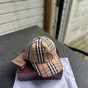 Burberry keps  - Snygg keps från Burberry med klassiskt rutigt mönster i beige, svart, vitt och röda linjer. Storlek M helt ny