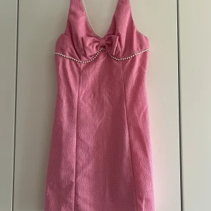 Rosa kort klänning med pärldetaljer - Supersöt rosa kort klänning med pärldetaljer längs bysten och en stor rosett framtill. Klänningen är ärmlös och har en lätt glittrig struktur i tyget, perfekt för dig som vill sticka ut. Snygg passform och v-ringad halsringning.