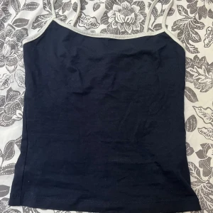 Mörkblå linnetopp från Bershka - Säljer en stilren mörkblå linnetopp från Bershka med tunna axelband och ljusblå kantdetaljer. Toppen är ärmlös och har en enkel, rak passform som är perfekt för varma dagar. Materialet är mjukt och bekvämt, vilket gör den lätt att bära hela dagen.