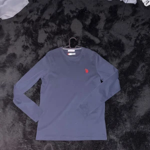  långärmad tröja US Polo Assn - Snygg marinblå långärmad tröja från US Polo Assn med röd broderad logga på bröstet. Tröjan har rund hals och är gjord i mjuk bomull, perfekt för dig som gillar stilrena och klassiska plagg med en sportig touch.