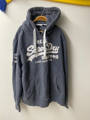 Mörkgrå Superdry hoodie med tryck - Säljer en mörkgrå hoodie från Superdry med stort vitt tryck på bröstet och siffror på ärmen. Tröjan har huva med breda snören och en klassisk känguruficka framtill. Perfekt för chill dagar och streetstyle.
