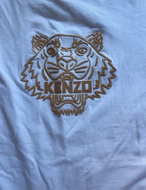 Vit Kenzo t-shirt med tigertryck - Säljer en vit t-shirt från Kenzo i bomull med ett snyggt guldigt tigertryck på bröstet. Klassisk rund hals och korta ärmar. Perfekt för dig som gillar streetstyle och vill sticka ut med ett ikoniskt motiv.