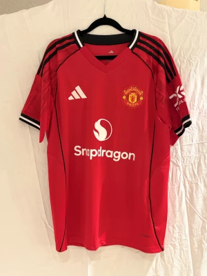 Manchester United Maguire #5 Adidas tröja - Röd Manchester United fotbollströja med Maguire #5 på ryggen, broderat klubbmärke och Adidas-logga på bröstet. Svarta och vita detaljer på ärmarna, samt sponsortryck. Tillverkad i lätt polyester med ventilerande meshpaneler.