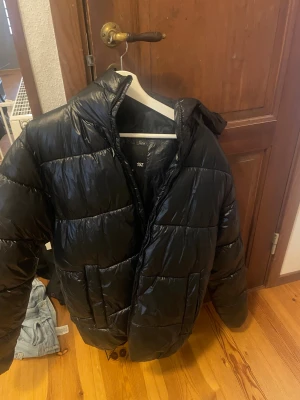 Svart pufferjacka - Svart pufferjacka från 157 i storlek S. Jackan har en glansig yta, hög krage och breda quiltade sektioner. Perfekt för kalla dagar och har en stilren look som funkar till många outfits. Dragkedja framtill och två stora fickor på sidorna.