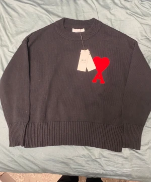 Svart stickad tröja från Ami - Svart stickad tröja från Ami med rund hals och ribbade muddar. På bröstet finns ett stort rött hjärta med bokstaven A, vilket ger en unik och trendig look. Tröjan är tillverkad i ekologisk bomull enligt GOTS-standard. Perfekt för dig som gillar stilrena detaljer.