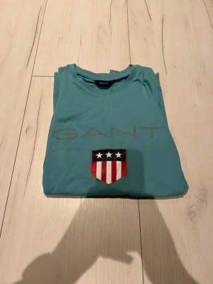 Turkos långärmad tröja från GANT - Snygg turkos långärmad tröja från GANT med stort tryck på bröstet och ett coolt emblem med stjärnor och rödvita ränder. Tröjan har rund hals och är gjord i mjuk bomull, perfekt för dig som gillar klassisk stil med en twist.