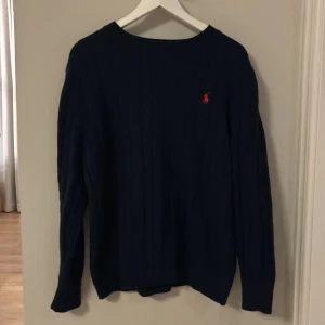 Mörkblå stickad tröja Polo Ralph Lauren - Snygg mörkblå stickad tröja från Polo Ralph Lauren i storlek m. Tröjan har klassiskt flätstickat mönster och en röd broderad logga på bröstet. Rund hals och långa ärmar, perfekt för dig som gillar preppy stil och vill ha något som känns både chill och stilrent.