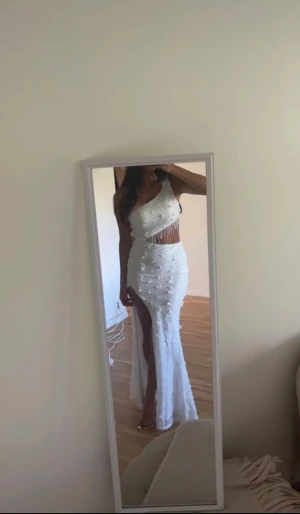 Vit långklänning med slits och pärlor - Supersnygg vit långklänning med oneshoulder och öppen rygg. Klänningen har glittriga pärldetaljer och fransar vid midjan, samt en hög slits på sidan. Materialet är glittrigt och har små blommor över hela klänningen. Perfekt för dig som vill sticka ut.