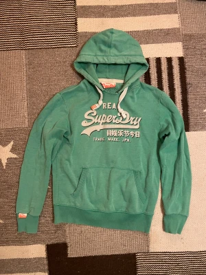 Superdry hoodie - Snygg grön hoodie från Superdry i storlek L med vit tryckt logga och japanska tecken på bröstet. Tröjan har huva med vita snören och ribbade muddar vid ärmslut och nederkant. Perfekt för dig som gillar streetwear och vill ha en chill look.