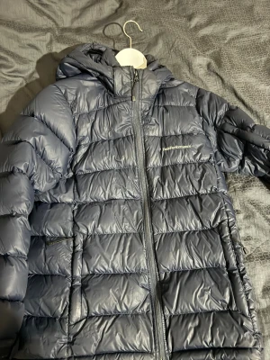 Mörkblå Peak Performance dunjacka - Säljer en mörkblå dunjacka från Peak Performance med huva och dragkedja framtill. Det finns en defekt i bak se på bild syns inte när man använder den, bra skick förutom de.