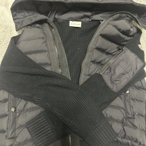 Svart cardigan från Moncler - Svart cardigan från Moncler med stickade ärmar och detaljer. Jackan har huva, dragkedja framtill och Moncler-logga på ärmen. Perfekt för dig som gillar streetstyle och vill ha något varmt och snyggt.
