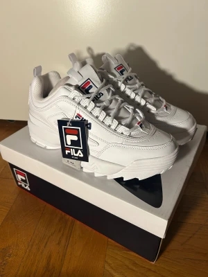 Fila Disruptor Premium vita sneakers - Fila Disruptor Premium sneakers i helvit med detaljer i marinblått och rött. Chunky design med platt sula och rund tå. Skorna har snörning och är tillverkade i syntetmaterial med Fila-logga på sidan och tungan. Perfekt för dig som gillar retrostil och streetwear. Strl i lager: 38, 39, 40.5 och 41.