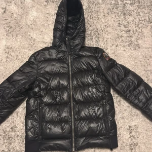 Svart pufferjacka från Parajumpers - Säljer en svart pufferjacka från Parajumpers, modell Sheen. Jackan har huva, dragkedja framtill och två sidofickor med dragkedja. Märkets patch sitter på vänster ärm. Jackan är quiltad och har en glansig finish.