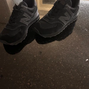 Svarta New Balance 574 sneakers - Säljer ett par svarta New Balance 574 sneakers med klassisk mesh och mocka på ovandelen. Skorna har en tydlig N-logga på sidan, snörning och en platt sula med bra grepp. Perfekt för dig som gillar stilrena och sportiga sneakers med retrovibe.