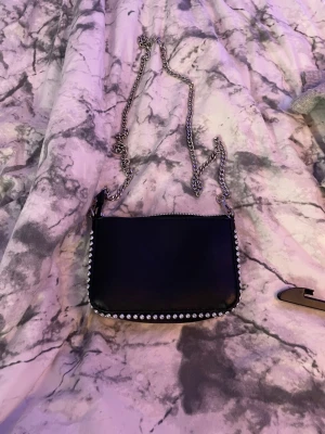 Svart axelväska med kedja och nitar - Snygg svart axelväska med silverfärgad kedja som axelrem och coola nitar längs kanterna. Väskan har en enkel rektangulär form och dragkedja upptill. Perfekt för att lyfta din outfit med lite edge och stil.