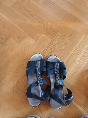 Svarta sandaler med strassdetaljer - Snygga svarta sandaler med breda elastiska band och en rem med glittrande strass framtill. Platt sula och öppen tå gör dem perfekta för varma dagar. Materialet är en mix av syntet och mocka-look. Passar dig som vill ha både stil och komfort i sommar.