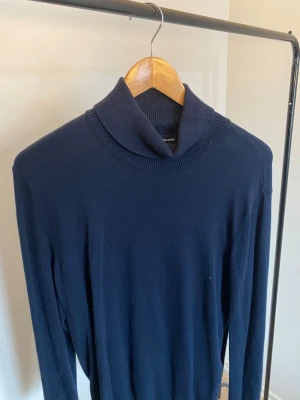 Mörkblå polotröja från Jack & Jones - Snygg mörkblå polotröja från Jack & Jones i mjuk stickad bomull. Tröjan har lång ärm och en bred, ribbad polokrage som ger en clean och stilren look. Perfekt för dig som gillar klassiska och enkla plagg med lite extra edge.