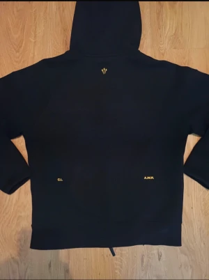 Svart Nike NOCTA hoodie med dragkedja - Svart hoodie från Nike NOCTA med gul logga på bröstet och NOCTA-tryck på ärmen. Tröjan har hel dragkedja, två fickor med dragkedja och stor huva. Materialet är mjukt och känns som polyester, perfekt för chill eller träning.