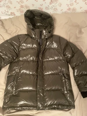  Super Puff olive green pufferjacka  - Säljer en mörkgrå pufferjacka från The Super Puff i storlek L. Jackan har glansig yta, stor huva med snörning och dragkedja framtill. Fylld med dun för extra värme och har två stora fickor med dragkedja. Perfekt för kalla dagar.