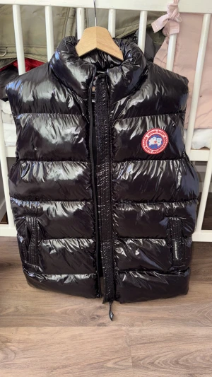 Svart dunväst från Canada Goose - Snygg svart dunväst från Canada Goose med glansig finish och klassisk logga på bröstet. Västen har hög krage, två fickor med dragkedja och quiltad design. Perfekt för lager-på-lager under kyliga dagar.