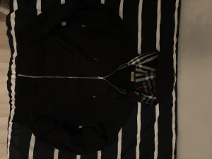 Svart Burberry hoodie med dragkedja - Svart hoodie från Burberry med klassiskt rutigt mönster i huvan och broderad logga på bröstet. Tröjan har hel dragkedja framtill, långa ärmar och huva. Materialet är mjukt och känns som bomull, perfekt för chill dagar. Är storlek L men passar även M. Hör av dig om du har några frågor