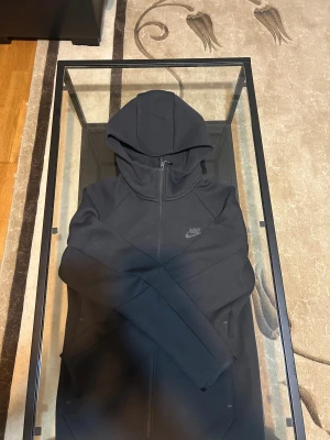 Svart hoodie från Nike med dragkedja - Svart hoodie från Nike i storlek XS med hel dragkedja framtill och huva. Tröjan har diskret Nike-logga på bröstet och är tillverkad i mjukt bomullsmaterial. Snygg och enkel design med långa ärmar och praktisk ficka med dragkedja på sidan av huvan. Helt ny