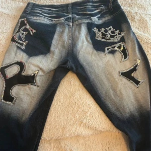 Blue Queen jeans med paljettdetaljer - Unika jeans från Blue Queen i blått med kraftig blekning och stora svarta bokstavs-applikationer med paljetter och en krona. Sönder på baksidan men inget man märker. Bootcut storlek 40! Passar mig perfekt som är ca 160! 