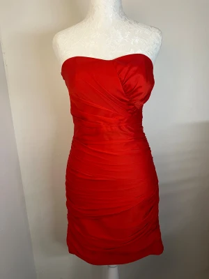 Klänning - Röd axelbandslös klänning med draperad design och guldig dragkedja baktill. Figurnära passform, storlek 36 (S) men är något mindre i storleken så känns mer som XS. Använd en gång och i utmärkt skick ❤️
