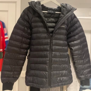 Svart dunjacka från Peak Performance - Säljer en svart dunjacka från Peak Performance med huva och två sidofickor med dragkedja. Jackan har quiltad design och Peak Performance-logga på bröstet samt en bred rem med logga på huvan. Nästan helt ny, använt bara någon gång. Direkt köpt från butik. Perfekt för kalla dagar och har en snygg, sportig look. Dam jacka storlek M