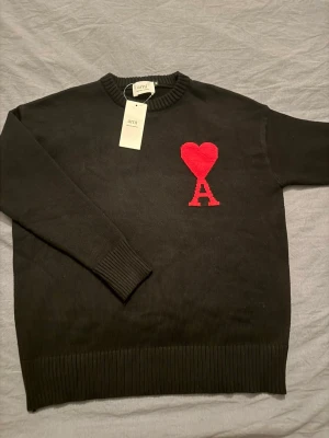 Svart stickad tröja från Ami - Svart stickad tröja från Ami med röd hjärt- och A-logga på bröstet. Rund hals och ribbade muddar vid ärmslut och nederkant. Snygg och stilren design som sticker ut med den ikoniska detaljen framtill.