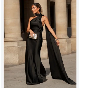 Svart långklänning med oneshoulder/ balklänning  - Säljer en elegant svart långklänning i satin med oneshoulder och lång scarf-detalj som faller ner från halsen. Klänningen har öppen rygg och en tight passform som framhäver siluetten. Perfekt för dig som vill sticka ut med en sofistikerad look. Perfekt som balklänning. Säljer då den inte passade mig