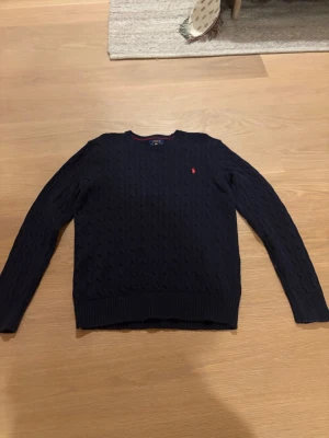 Mörkblå stickad tröja Polo Ralph Lauren - Snygg mörkblå stickad tröja från Polo Ralph Lauren i storlek XL. Tröjan har klassiskt kabelstickat mönster, rund hals och en röd broderad logga på bröstet. Perfekt för dig som gillar stilrena och tidlösa plagg med premiumkänsla.
