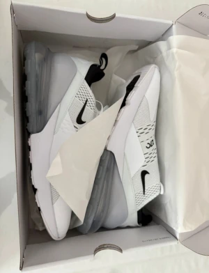 Nike Air Max 270 vita sneakers - Nike Air Max 270 sneakers i vitt med svarta detaljer och stor synlig luftbubbla i sulan. Ovandelen är i mesh och syntet för extra andningsförmåga och komfort. Snygg, sportig design med snörning och tydlig Air 270-logga på sidan och plösen.